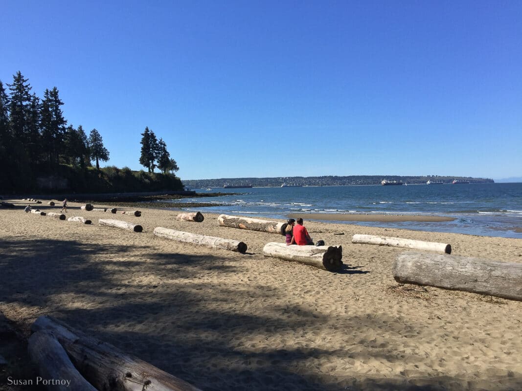 A couple sits on a log looking atA couple sits on a log looking at the water at Vancouver's Third Beach